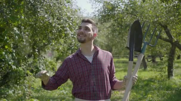 Jovem agricultor barbudo sorridente caminhando pelo jardim com uma pá e forquilha nas mãos. Conceito de vida rural, fruticultura, jardinagem. Movimento lento — Vídeo de Stock