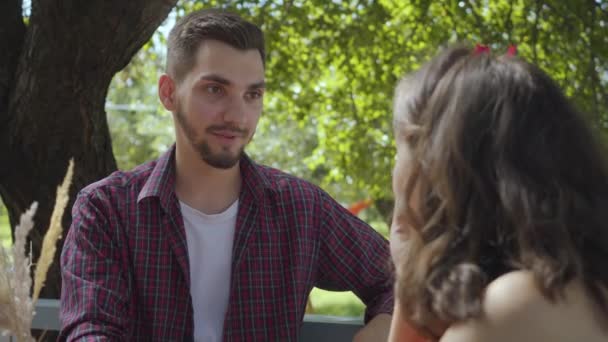 Retrato de cerca de una joven pareja feliz sentada al aire libre en la mesa hablando amistosamente y sonriendo entre sí. Hombre y mujer positivos relajándose en el patio trasero. Ocio en casa — Vídeos de Stock