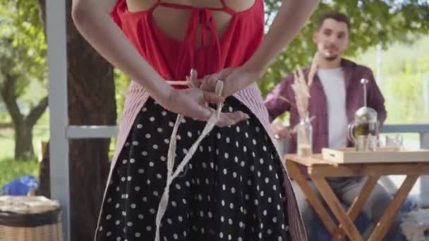 Vista posterior de la joven mujer en blusa retro y falda atando un delantal en primer plano mientras su marido se sienta a la mesa en el fondo. Feliz pareja vintage al aire libre — Vídeo de stock