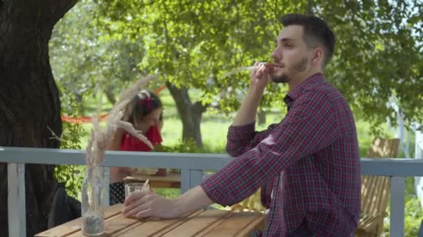 Unga skäggiga man dricka te i förgrunden medan koncentrerad attraktiv flicka i retro kläder plantera blommor i krukor i bakgrunden. Rutinmässig livslängd. Make avkopplande medan hustru arbetar — Stockvideo