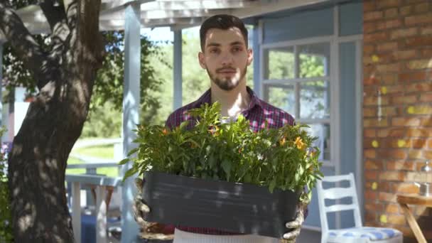 Retrato de un joven granjero barbudo sonriente sosteniendo una maceta de flores en las manos mirando a la cámara sonriendo de pie frente a la vieja casa. Concepto de vida rural, cultivo, jardinería. Serie de personas reales . — Vídeos de Stock