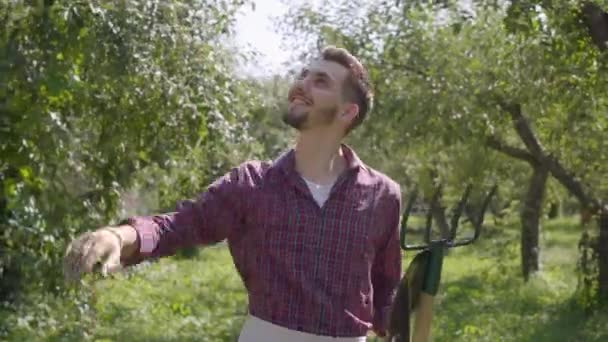 Joven y confiado agricultor caucásico barbudo sonriente caminando por el jardín con una pala y una horquilla revisando árboles frutales. Concepto de vida rural, fruticultura, jardinería . — Vídeos de Stock
