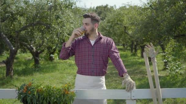 Joven agricultor confiado en guantes de jardín bebiendo el té de pie en el verde jardín de verano cerca de la cerca. El hombre descansando después del trabajo. Estilo de vida rural. Serie de personas reales . — Vídeos de Stock