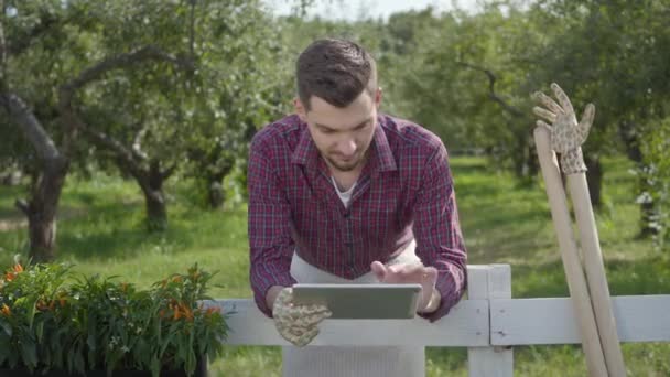 Молодий фермер використовує планшет, що стоїть за парканом на передньому плані, літній сад на задньому плані. Чоловік відпочиває після роботи. Сільський спосіб життя — стокове відео