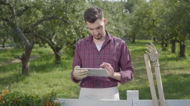 Jardineiro experiente, bem sucedido e profissional usando o tablet em pé atrás da cerca em primeiro plano, o jardim de verão em segundo plano. O homem que descansa depois do trabalho. Estilo de vida rural — Vídeo de Stock