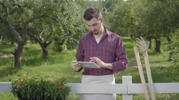 정원 장갑을 끼고 있는 젊은 농부는 전경에 있는 울타리 뒤에 서 있는 태블릿을 사용 하 여, 여름에는 여름 정원을 배경으로 합니다. 엄지 손가락을 보여주는 긍정적 인 남자. 농촌 생활 — 비디오