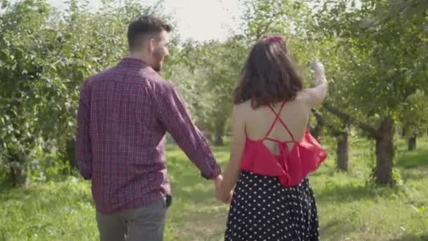 Vista posterior de la joven pareja positiva caminando por el jardín tomados de la mano y hablando. Feliz hombre y mujer pasando tiempo al aire libre. Conexión con la naturaleza, la vida rural, cita romántica. Vista trasera — Vídeos de Stock