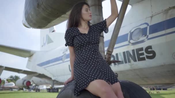 Ευτυχισμένο νεαρό μελαχρινή κορίτσι με μαύρο φόρεμα κοιτάζοντας την κάμερα κάθεται στον εξοπλισμό προσγείωσης του λευκού αεροπλάνου. Καλοκαίρι. Η χαρά του ταξιδιού. Η έννοια του ταξιδιού, αεροσκάφος, το Σαββατοκύριακο. Αργή κίνηση — Αρχείο Βίντεο