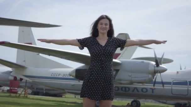 Happy young brunette girl in short dress looking at the camera leans to the side with arms apart, imitating plane in front of airplanes. Joy of travel. Concept of traveling, aircraft. Slow motion — Stock Video