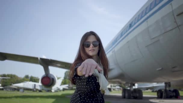 Portrait happy playful brunette girl wearing sunglasses in front of the big plane and looking at camera. Summertime. Joy of travel. Concept of traveling, aircraft, weekend. Slow motion — Stock Video
