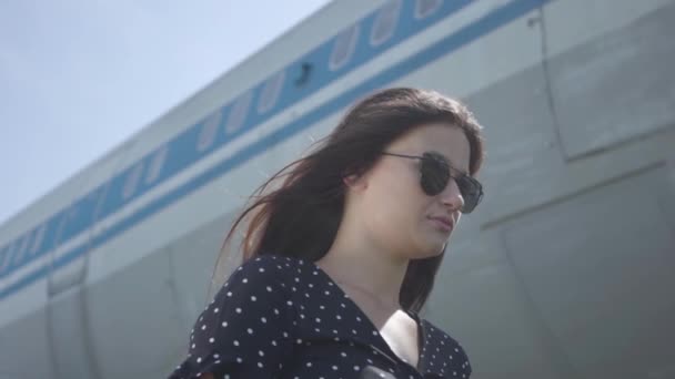 Portrait young confident brunette girl wearing sunglasses standing in front of the big plane. Summertime. Joy of travel. Concept of traveling, aircraft, weekend. Slow motion — Stock Video