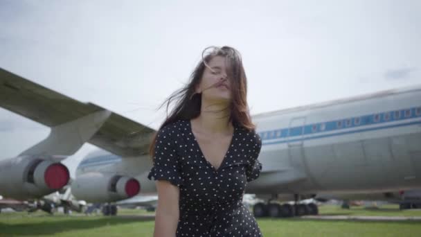Όμορφη ευτυχισμένη μελαχρινή κορίτσι χαμογελά και κοιτάζοντας την κάμερα μπροστά από το μεγάλο λευκό αεροπλάνο, τα μαλλιά φτερουγίζουν στον άνεμο. Γυναίκα που περπατάει στο αεροπλάνο. Η χαρά του ταξιδιού. Ταξίδια Concept, αεροσκάφη — Αρχείο Βίντεο