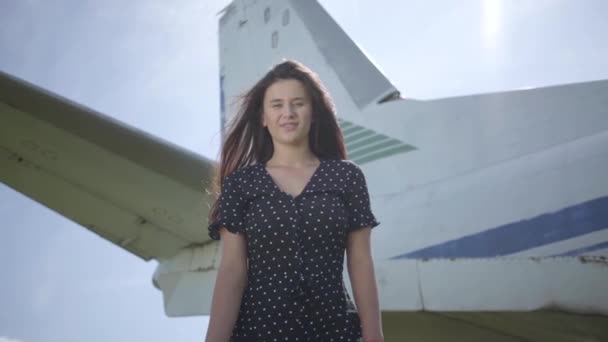 Šťastná mladá usměvavá brunetka se dívá na kameru před velkým bílým letadlem. Létě. Radost z cestování. Koncept cestování, letadel, víkend. Zpomaleně — Stock video