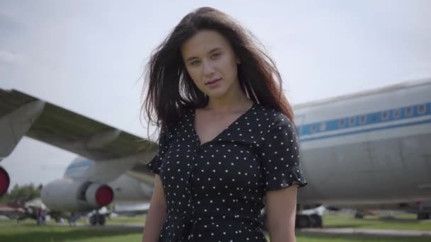 Joyeux jeune fille brune dansant et tournant autour en regardant la caméra devant le grand avion blanc, les cheveux flottant sur le vent. L'été. Joie du voyage. Concept de voyage, avion — Video