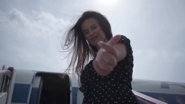 Lycklig ung brunett tjej med fladdrande hår gör inbjudande gest tittar på kameran framför planet. Rese glädjen. Koncept för resande, flygplan — Stockvideo
