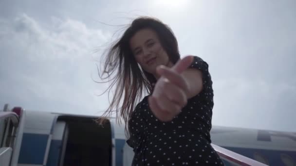 Menina morena feliz com cabelo agitado fazendo gesto convidativo olhando para a câmera na frente do avião. Alegria da viagem. Conceito de viajar, avião — Vídeo de Stock