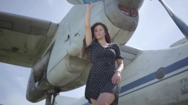 Jeune fille brune confiante vêtue d'une robe noire regardant la caméra en face de l'avion blanc toucher. Joie du voyage. Concept de voyage, avion, week-end. Mouvement lent . — Video