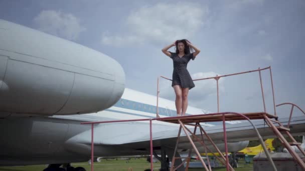 Счастливая молодая улыбающаяся брюнетка смотрит в камеру, стоя на большой белый самолет, трогающий ее волосы. Летом. Радость путешествий. Концепция путешествия, самолеты, выходные. Медленное движение — стоковое видео