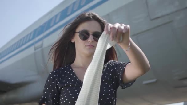 Ritratto bella ragazza bruna con lo scialle bianco e occhiali da sole di fronte al grande aereo e guardando la fotocamera. Ora legale. Gioia del viaggio. Concetto di viaggio, aereo, fine settimana. Rallentatore — Video Stock