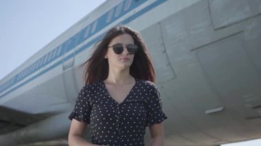 Portre sevimli genç kendinden emin esmer kız büyük uçağın önünde duran güneş gözlüğü giyiyor. Yaz. Seyahat insebunu. Seyahat kavramı, uçak, hafta sonu. Yavaş çekim