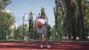 Sevimli genç esmer kız basketbol sahası açık havada duran kameraya bakarak bir basketbol topu tutan. Spor kavramı, güç, rekabet, aktif yaşam tarzı. Spor ve rekreasyon.