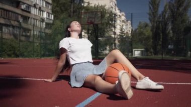 Basketbol sahası açık havada oturan kameraya bakarak bir basketbol topu ile sevimli genç esmer kız. Spor kavramı, güç, rekabet, aktif yaşam tarzı. Spor ve rekreasyon.