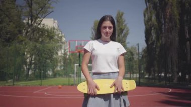 Açık havada basketbol sahası üzerinde duran kameraya bakan bir kaykay ile sevimli kadın portresi. Spor kavramı, rekabet, aktif yaşam tarzı. Spor ve rekreasyon.