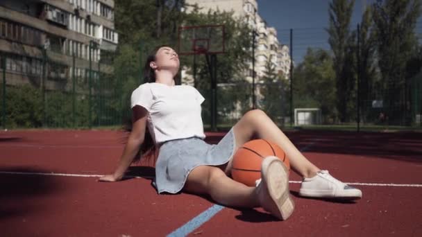 Мила дівчина брюнетка з баскетбольним м'ячем дивиться на камеру, що сидить на баскетбольному майданчику на відкритому повітрі. Концепція спорту, влади, конкуренції, активного способу життя. Спорт і відпочинок . — стокове відео
