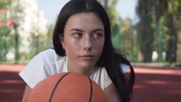 カメラを見てバスケットボールボールを持つ魅力的なブルネットの女性のクローズアップ顔。スポーツ、パワー、競争、アクティブなライフスタイルの概念。コートでバスケットボールをしている少女 — ストック動画