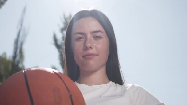 Porträt des netten brünetten Mädchens mit einer Zahnspange, die einen Basketballball hält und lächelnd in die Kamera schaut. Konzept von Sport, Macht, Wettbewerb, aktivem Lebensstil. Mädchen spielt Basketball auf dem Platz — Stockvideo