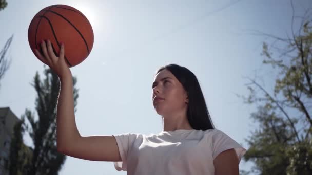 Porträtt av söt ung brunett kvinna kastar bollen upp. Begreppet idrott, makt, konkurrens, aktiv livsstil. Flickan spelar basket på spelplanen. Nedersta vyn. Sport och fritid. — Stockvideo