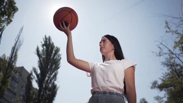 ボールを投げるかわいいブルネットの女の子の肖像画。スポーツ、パワー、競争、アクティブなライフスタイルの概念。コートでバスケットボールをしている少女。下のビュー。スポーツとレクリエーション. — ストック動画