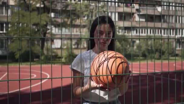 Retrato de adolescente morena con pelota de baloncesto mirando a la cámara detrás de la valla de malla en la cancha de baloncesto. Concepto de deporte, competición, estilo de vida activo. Deportes y recreación — Vídeo de stock