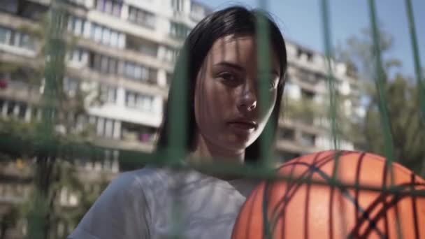 Portrait d'une fille brune tenant une balle de basket regardant la caméra debout derrière la clôture en filet du terrain de basket-ball. Concept de sport, compétition, mode de vie actif. Sports et loisirs . — Video