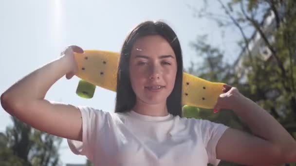 Retrato de una hermosa joven con un monopatín mirando a la cámara de pie al aire libre. Concepto de deporte, competición, estilo de vida activo. Deportes y recreación . — Vídeo de stock