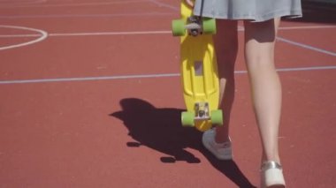 Basketbol sahasında yürüyen sarı bir kaykay ile kısa etek bacaklar kadın. Spor ayakkabılı kadın bacakları sarı kaykay ala. Gölge kızı takip ediyor. Spor kavramı, aktif yaşam tarzı