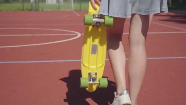 Láb a nő rövid szoknya egy sárga gördeszka séta a kosárlabdapálya. Shadow követi a lányt. A sport, a hatalom, a versengés, az aktív életmód fogalma — Stock videók