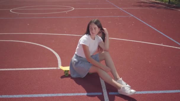 Menina morena bonito vestido com tênis, uma saia e uma camiseta senta-se em um skate na quadra de basquete ao ar livre em tempo de verão ensolarado. Conceito de esporte, estilo de vida ativo. Esportes e recreação . — Vídeo de Stock