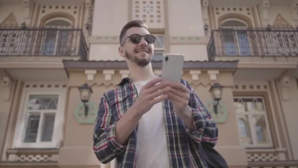 Jeune homme barbu en chemise à carreaux et lunettes de soleil regarde autour tout en étudiant l'architecture de la ville. Un touriste parcourt les rues de la ville. Tourisme et concept de voyage . — Video