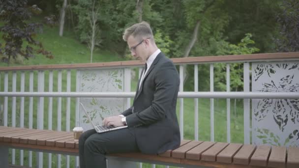 Jeune homme d'affaires attrayant et confiant en costume officiel travaillant avec un ordinateur portable et buvant du café dans le parc. Concept d'entreprise, freelance . — Video