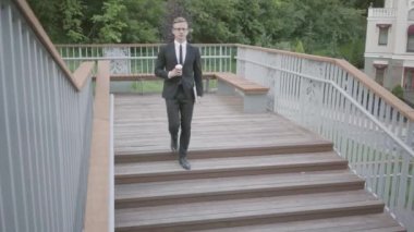 Genç iyi giyimli işadamı merdivenlerden inip elinde bir bardak kahve yle yürüyor. Ofis yaşam tarzı, iş konsepti. Parkta yürüyen takım elbiseli adam. Merdivenlerden aşağı