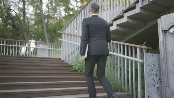 Successful stylish well-dressed businessman in expensive suit and shoes walking upstairs. Office lifestyle, business concept. The man in the suit holding a laptop walking outdoors. Climbing stairs. — Stock Video