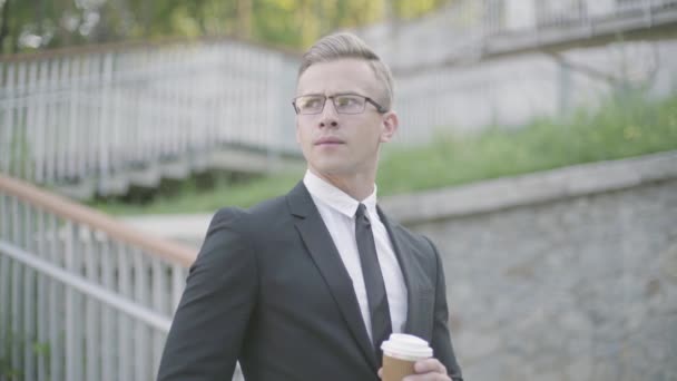 コーヒーを飲みながら階段の前に立つメガネをかけたハンサムな自信に満ちた若いビジネスマンの肖像画。屋外でパートナーを待っているオフィスマネージャー. — ストック動画