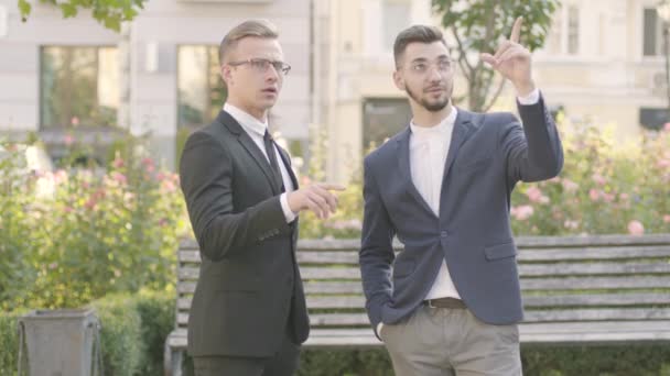 Two young well-dressed partners have meeting in the park. Confident businessman discussing with architect the project of new buildings outdoors. Business meeting, business relationship, property — Stock Video