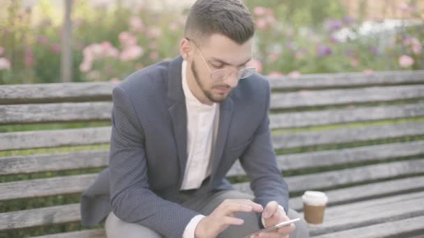 Портрет молодого бізнесмена, що сидить на лавці на вулиці. Дивлячись на його смартфон і набираючи повідомлення. Класично одягнений. Виглядати успішно, впевнено . — стокове відео