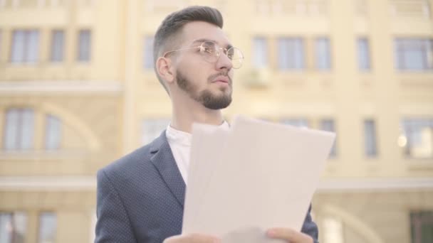 Porträt eines jungen, gut gekleideten Geschäftsmannes mit Brille, der vor hohen Gebäuden steht und Zeitungen liest. der Büroleiter, der im Freien arbeitet. Büro-Lifestyle, Geschäftskonzept — Stockvideo