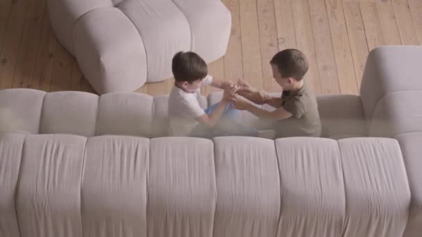 Δύο αξιαγάπητα αδέλφια που γελάνε και μαλώνουν μεταξύ τους στον καναπέ. Τα αδέλφια περνούν χρόνο στο σπίτι. Τα παιδιά τριγυρίζεις μέσα σε εσωτερικούς χώρους. Ευτυχισμένη οικογένεια, ανέμελη παιδική ηλικία. Κορυφαία προβολή — Αρχείο Βίντεο