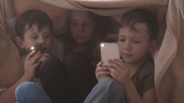 Två bröder och syster sitter under täcket leker med mobiltelefoner tillsammans. Syskon spenderar tid hemma. Lycklig familj, bekymmerslös barndom — Stockvideo