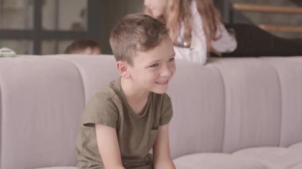 前景のソファに座って笑う小さな男の子の肖像画。バックグラウンドで遊んでいる姉妹と兄弟。兄弟関係、気楽な子供時代 — ストック動画