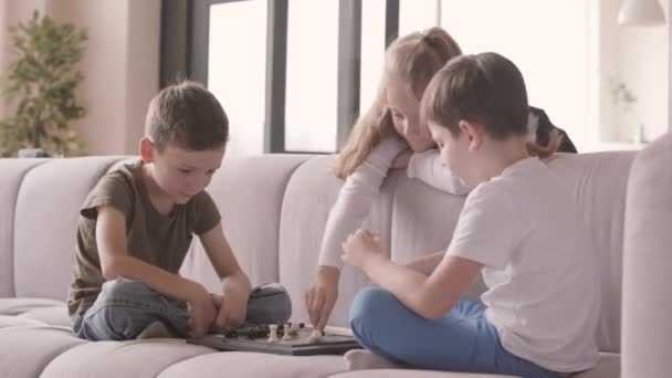 Twee jongens spelen Schaken zittend op de Bank thuis terwijl oudere zus kijken ze. Broers en zussen tijd binnenshuis doorbrengen. Gelukkige familie, zorgeloze kindertijd. Intelligent spel — Stockvideo